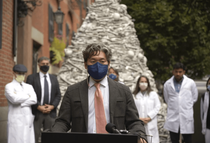 Dr. KJ Seung, a professor at Harvard Medical School, speaks at a September rally outside the home of the CEO of Moderna, maker of one of the two mRNA vaccines that prevent COVID-19. The fake bones are meant to represent lives lost unnecessarily to the coronavirus. Seung was part of a group of doctors demanding Moderna share vaccines — and its recipe — with low-resource countries. Credit: Jasper G. Goodman/Harvard Crimson